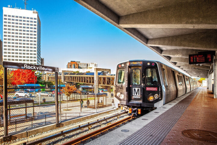 A Metro train arrives at the outdoor Rockville station platform under clear skies, with buildings and autumn trees visible in the background.