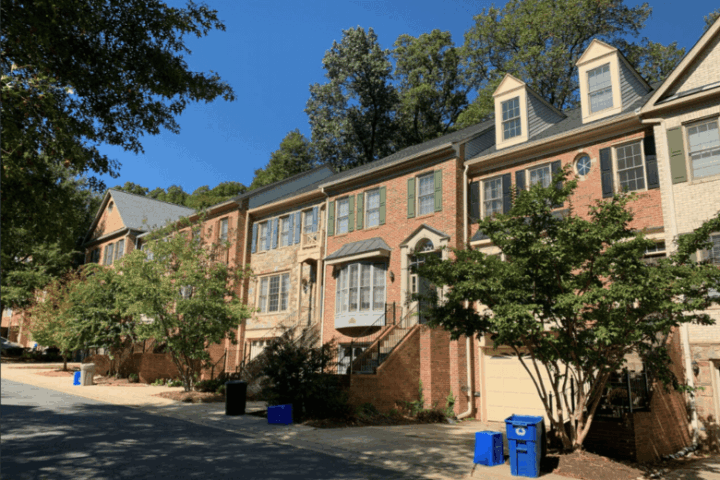 Row of multi-story brick townhouses with trees in front, blue recycling bins near the curb, and clear blue sky above.