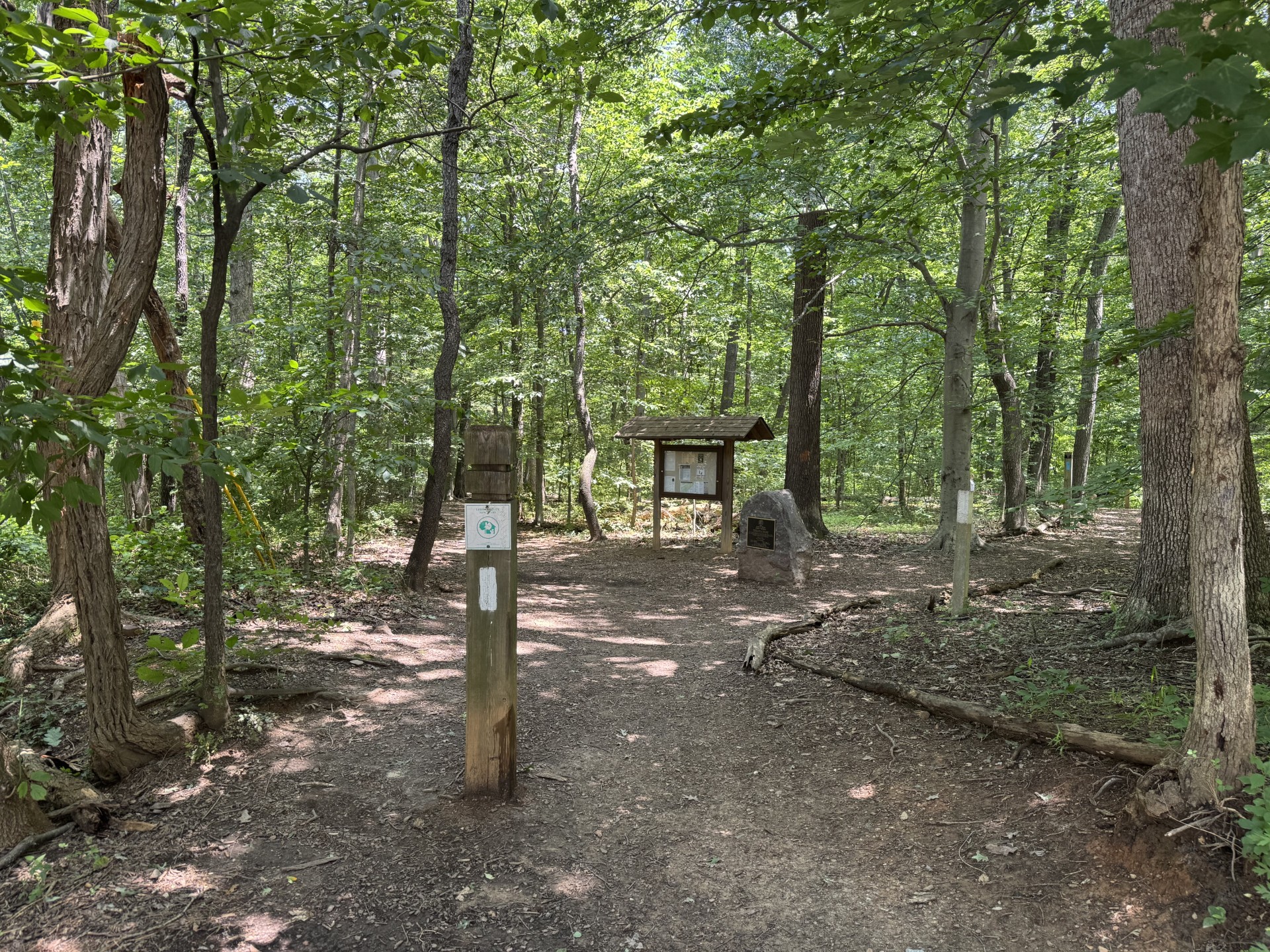 A wooded trailhead with a signpost, a map board under a shelter, and a large rock surrounded by leafy trees.