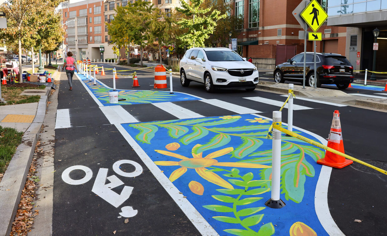 A city street features painted bike lanes with colorful floral designs, white crosswalk markings, traffic cones, and cars stopped at a pedestrian crossing.