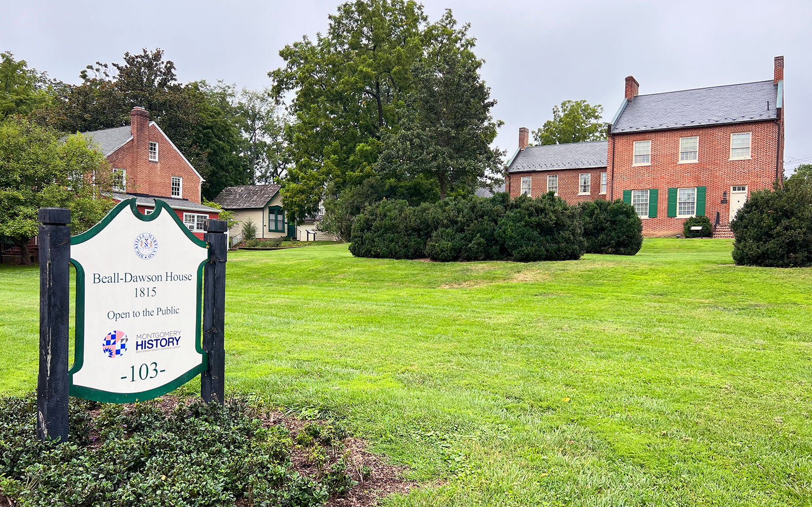 A brick house with green shutters sits on a grassy lawn; a sign in front reads "Beall-Dawson House 1815, Open to the Public," with symbols for history and the number 103.
