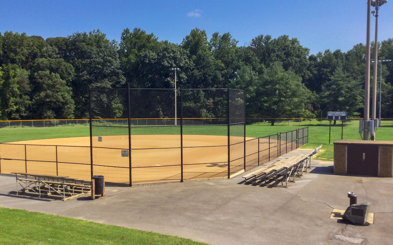 Empty baseball field with a dirt infield, metal bleachers, a chain-link fence, and green trees in the background under a clear blue sky.