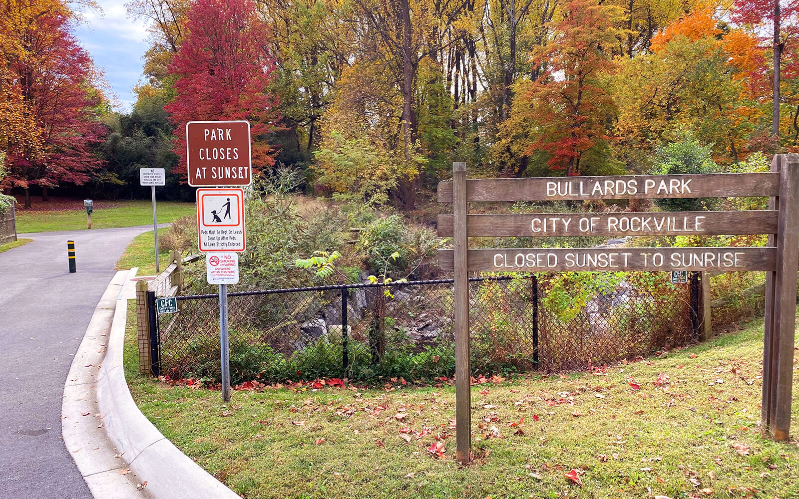 Entrance to Bullards Park in Rockville with signs stating the park closes at sunset and is closed from sunset to sunrise; trees with autumn foliage in the background.