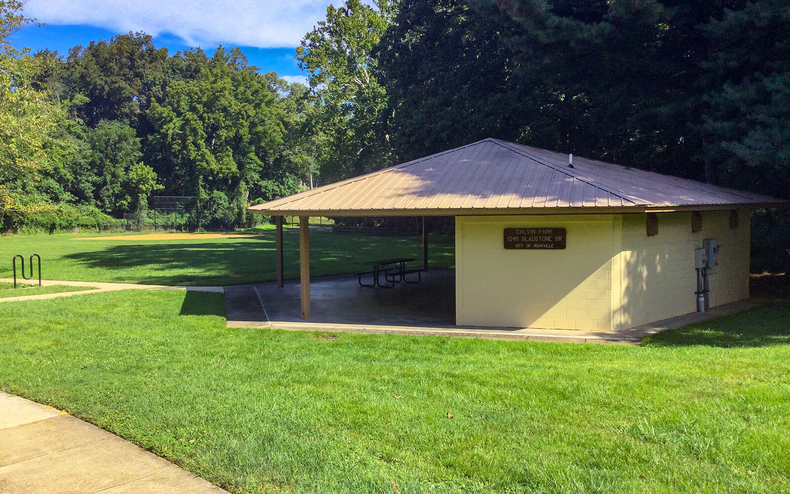 A small pavilion with picnic tables stands on a concrete pad in a grassy park area, surrounded by trees under a partly cloudy sky.