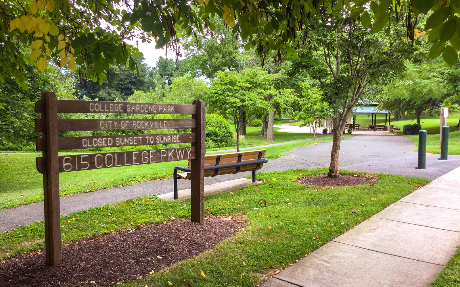 Wooden sign for College Gardens Park with park benches, paved path, trees, and a shelter in the background on a sunny day.