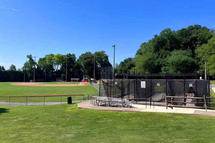 A baseball field with a green outfield, infield, and dugouts is shown next to batting cages surrounded by fencing and benches, with trees and blue sky in the background.