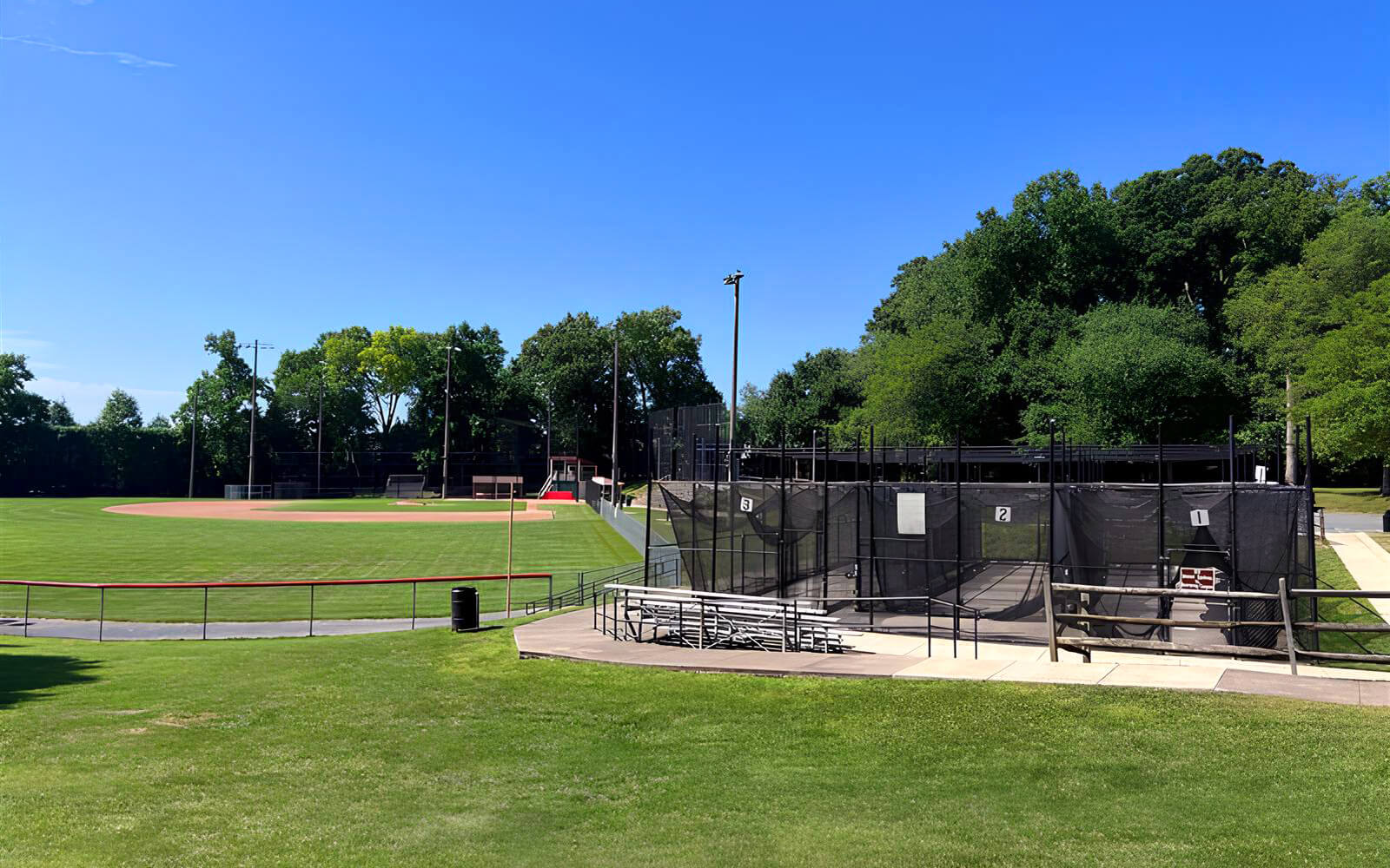 A baseball field with a green outfield, infield, and dugouts is shown next to batting cages surrounded by fencing and benches, with trees and blue sky in the background.