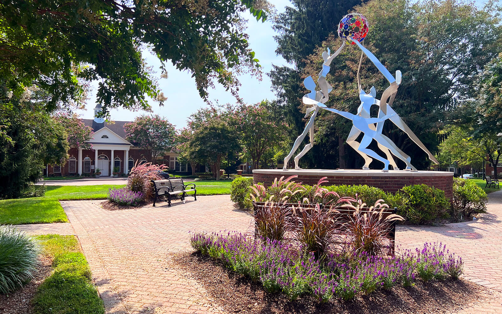 A metal sculpture of abstract human figures reaching for a colorful sphere stands in a landscaped brick courtyard with benches, flowers, and trees near a traditional building.