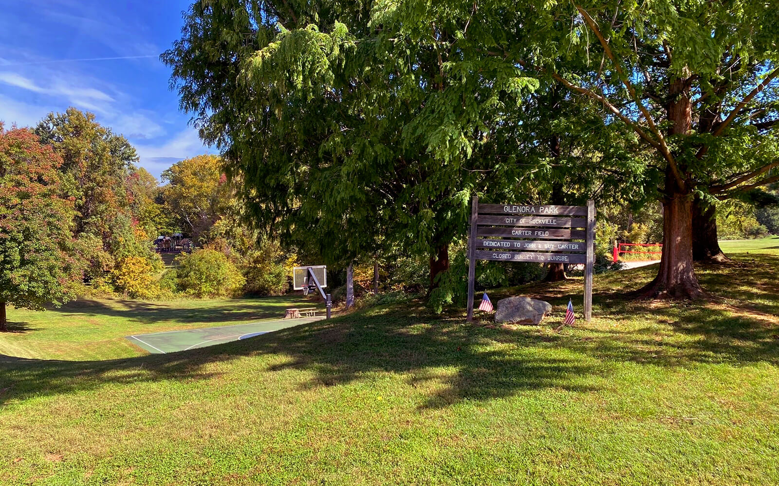 A grassy park area with trees, a basketball court, and a wooden sign displaying information near the entrance on a sunny day.