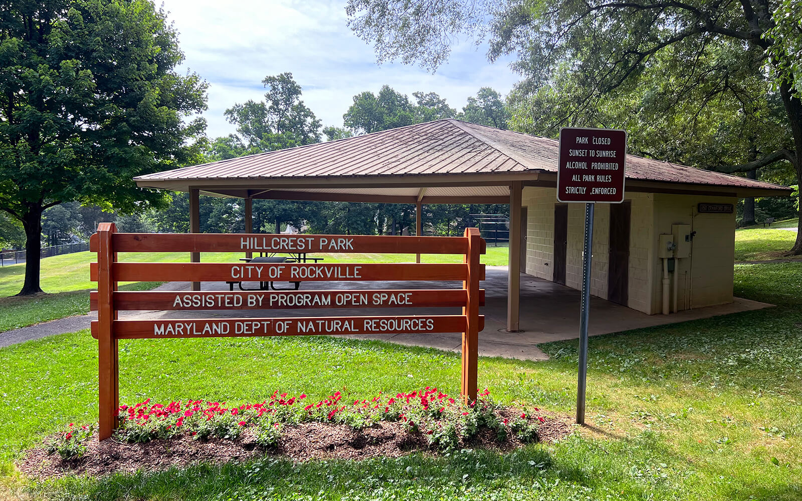 A wooden sign for Hillcrest Park stands in front of a pavilion and trees. The sign notes assistance from open space programs and Maryland Dept. of Natural Resources.