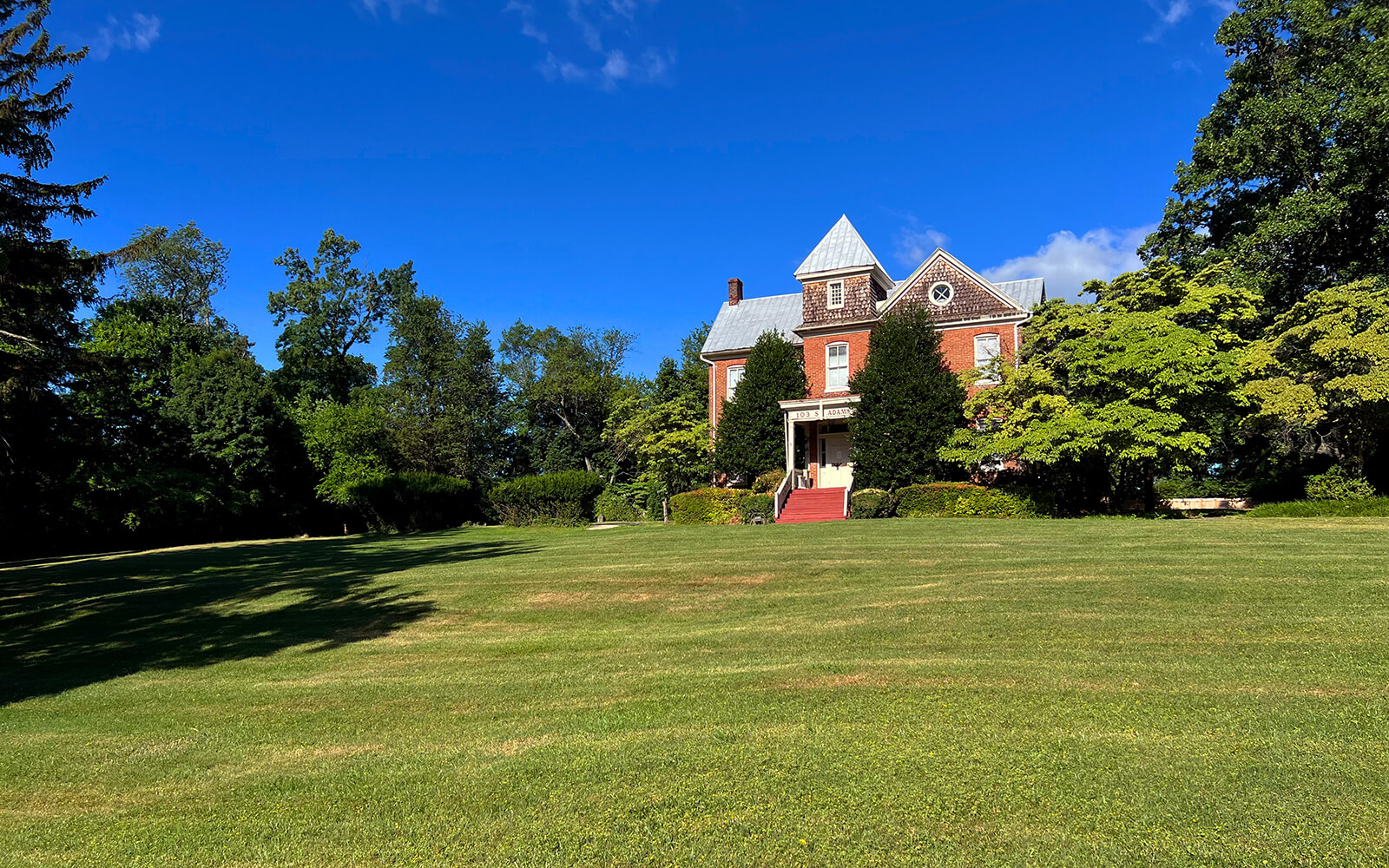 A large brick house with a peaked roof stands at the edge of a wide, neatly mowed lawn, surrounded by green trees under a clear blue sky.