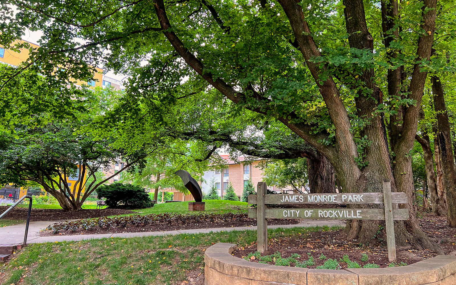 James Monroe Park entrance with a wooden sign under large trees, a pathway, and a sculpture visible in the background in Rockville.