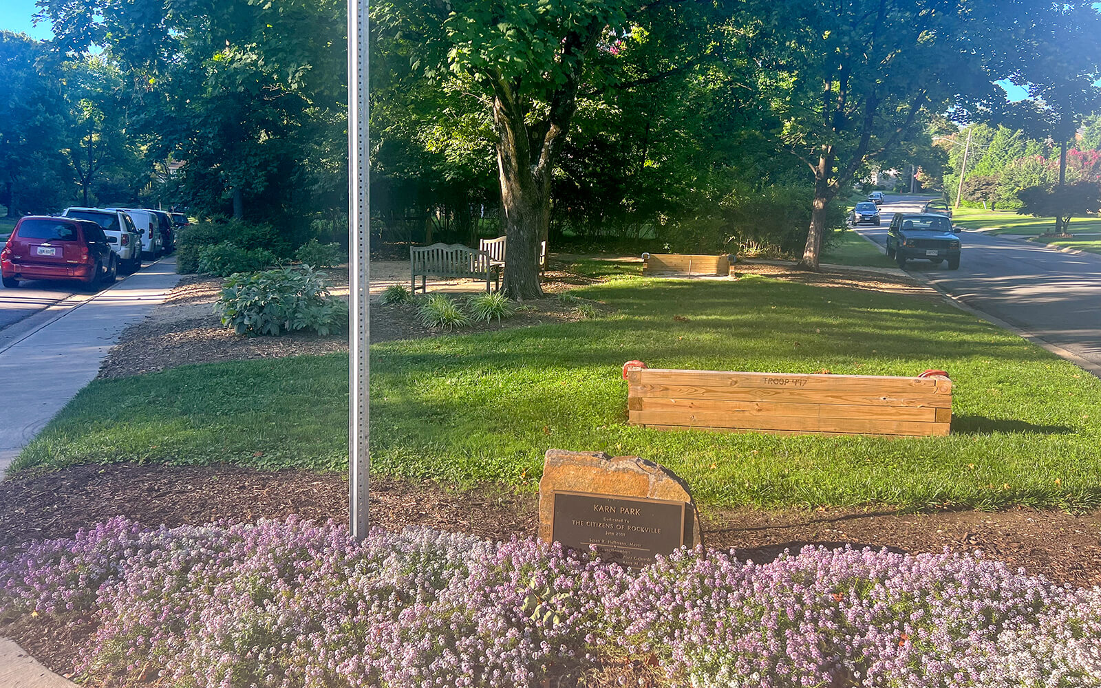 Small park area with a bench, a flower bed, a stone plaque, a wooden planter box, trees, parked cars, and a street visible on a sunny day.