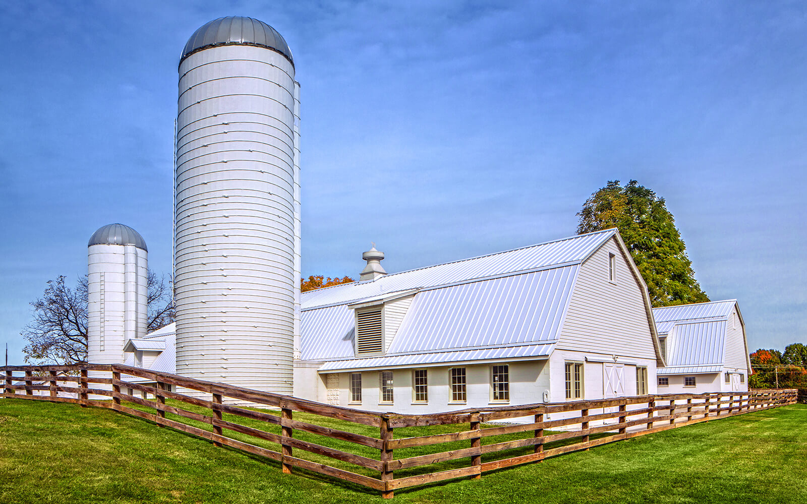 A white barn with two tall silos and a wooden fence sits on a grassy field under a blue sky.