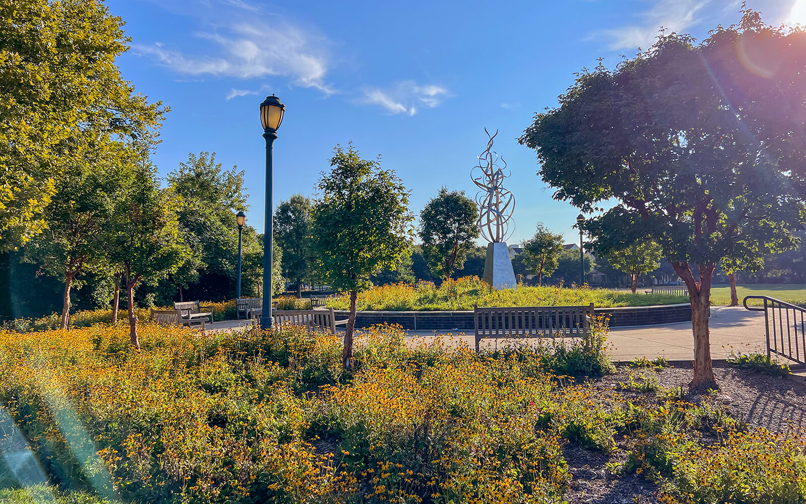 Sunny park scene with green trees, benches, a lamppost, gardens, and a tall abstract metal sculpture on a pedestal in the background.