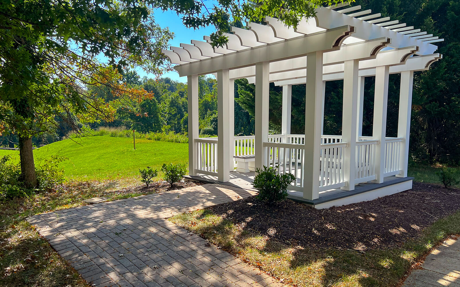 A white wooden pergola with railings stands on a mulched area next to a brick walkway, surrounded by green grass and trees under a sunny sky.