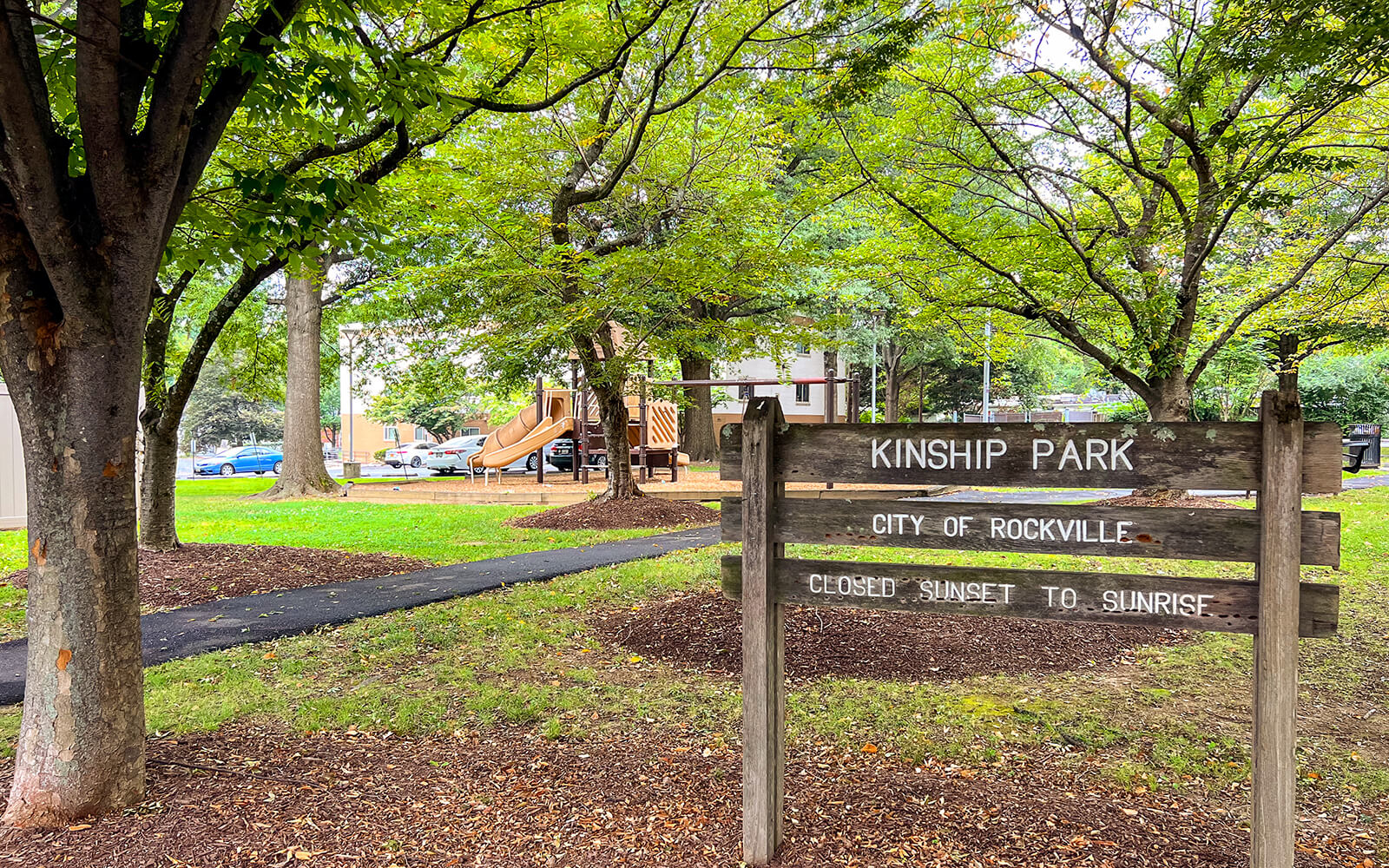 A wooden sign reads "KINSHIP PARK CITY OF ROCKVILLE CLOSED SUNSET TO SUNRISE" in a park with trees, a playground, and a paved pathway visible in the background.