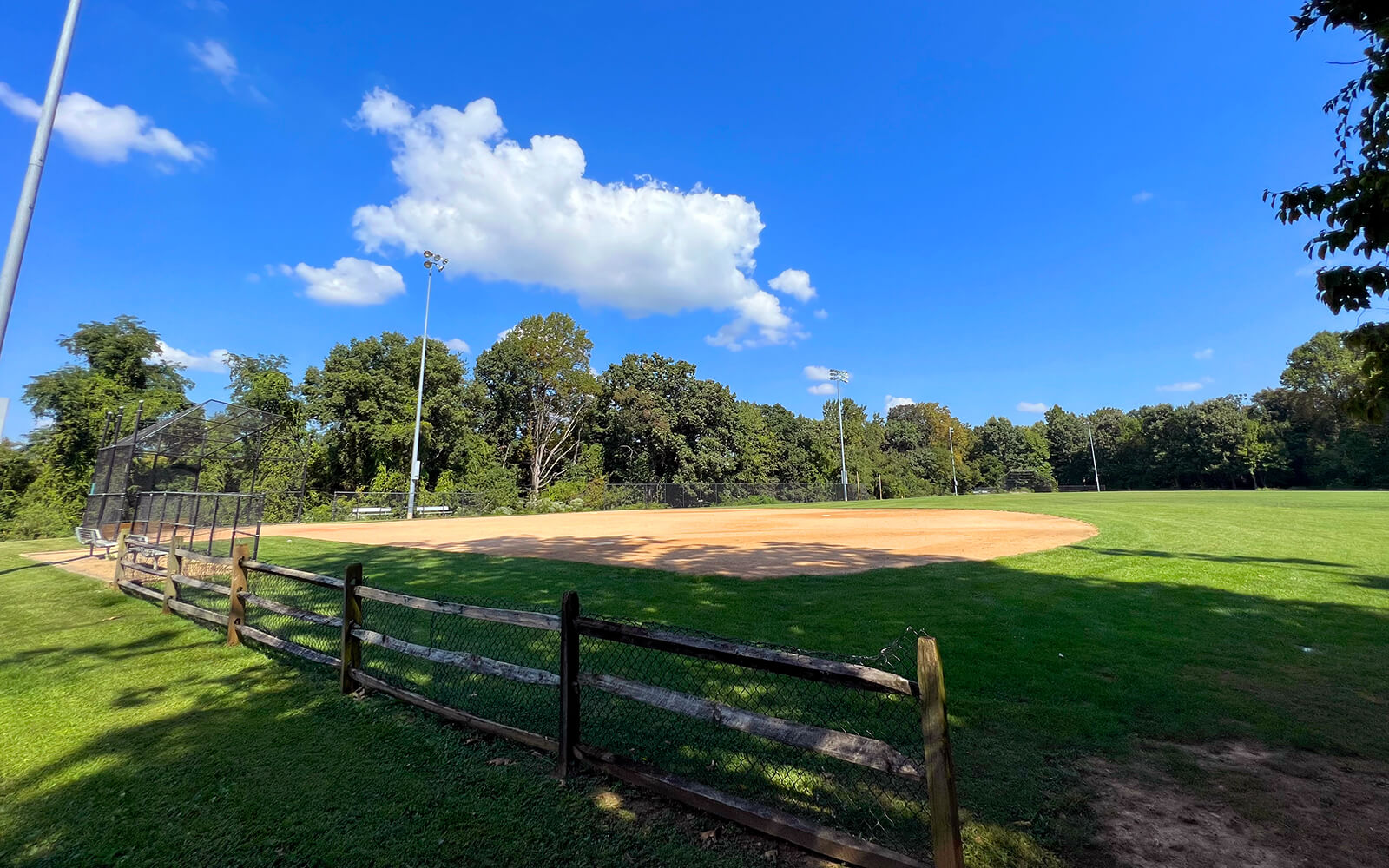 A baseball field with a dirt infield, green grass, and a wooden fence in the foreground, surrounded by trees under a blue sky with scattered clouds.