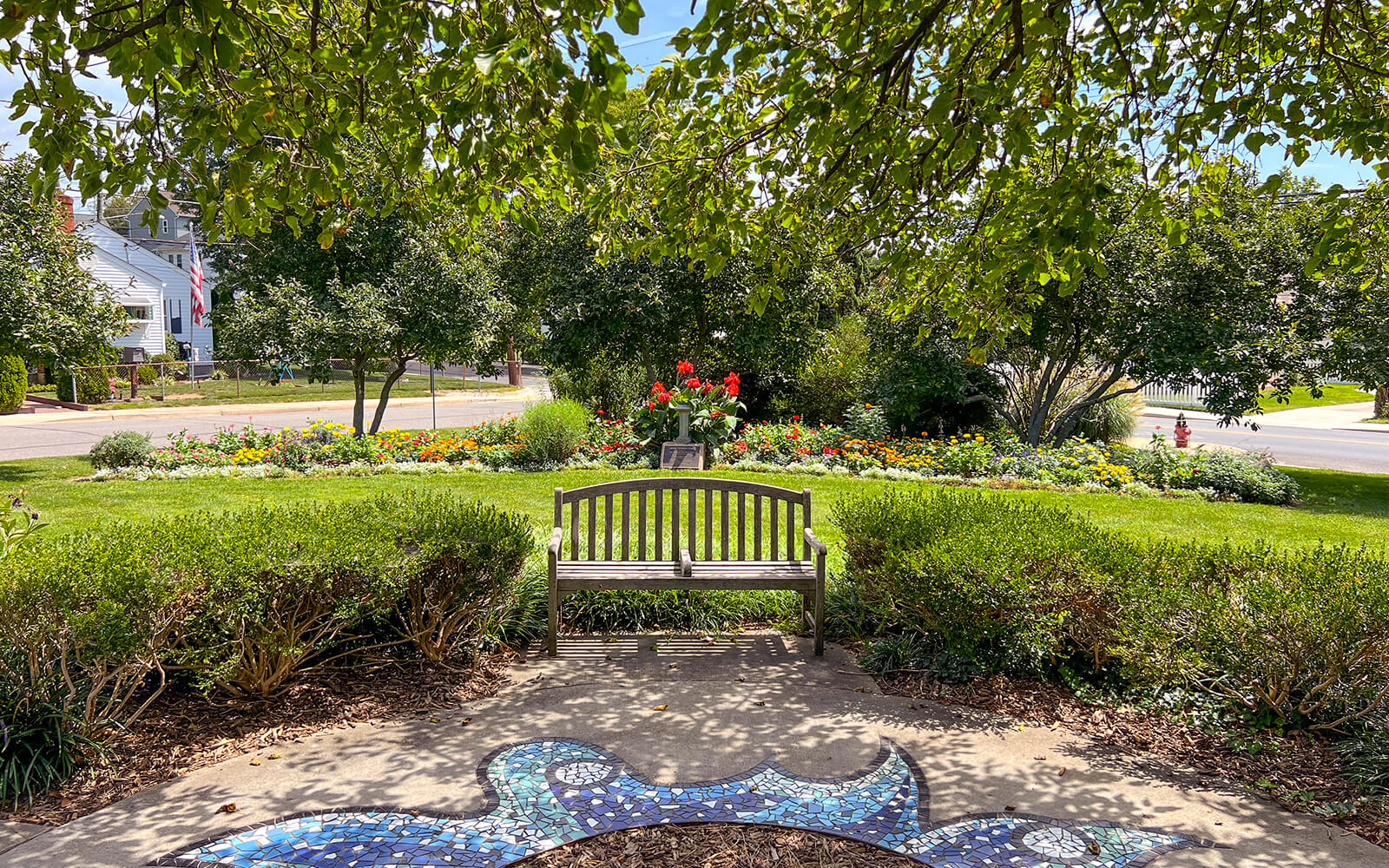 A wooden bench faces a flower garden bordered by shrubs and trees, with a mosaic design on the walkway in the foreground.