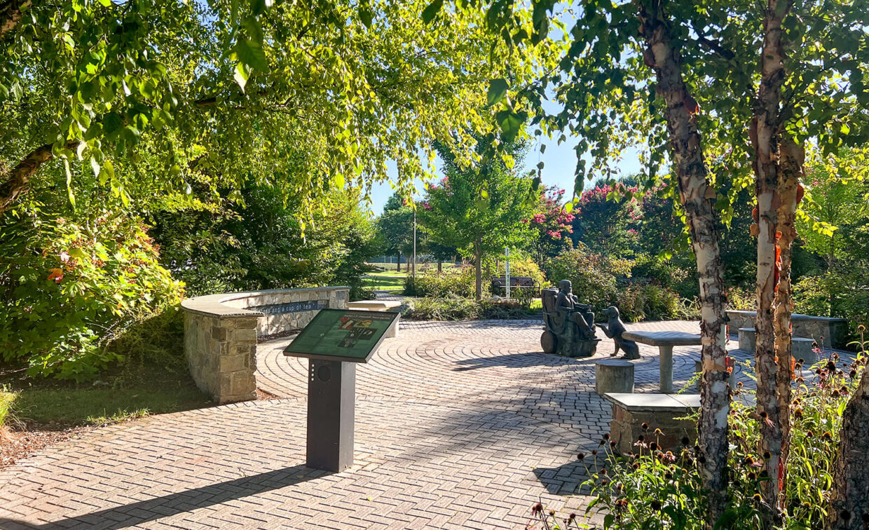 A sunlit park scene with a curved stone bench, an informational sign, and a bronze sculpture of people sitting on benches surrounded by trees and greenery.