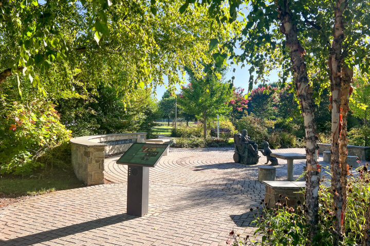 A sunlit park scene with a curved stone bench, an informational sign, and a bronze sculpture of people sitting on benches surrounded by trees and greenery.