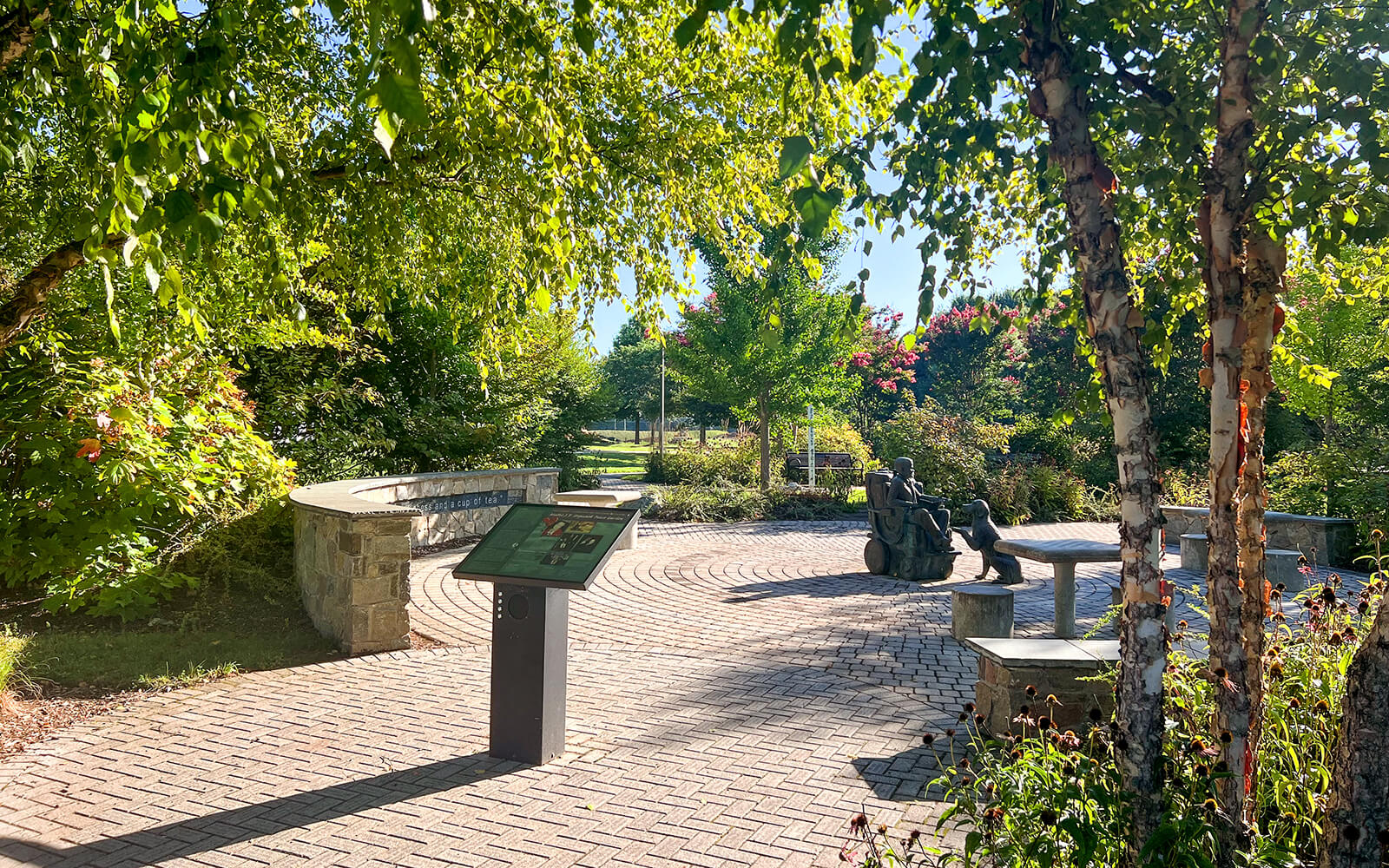 A sunlit park scene with a curved stone bench, an informational sign, and a bronze sculpture of people sitting on benches surrounded by trees and greenery.