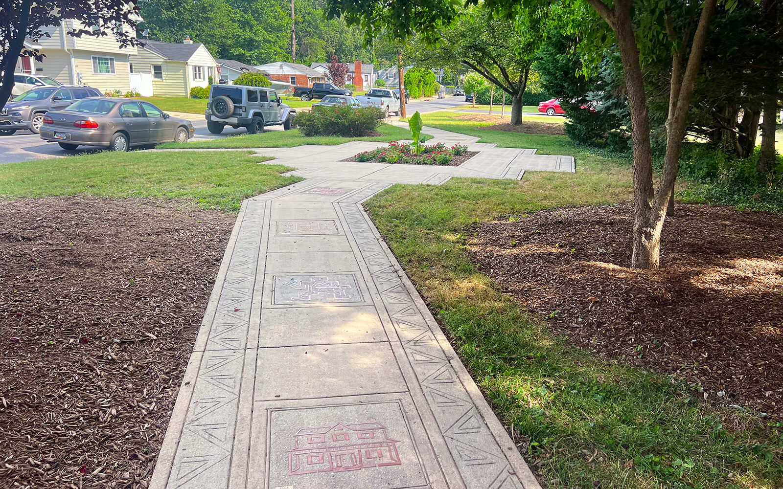 A paved sidewalk with geometric patterns runs through a grassy area with trees, leading towards a residential street with parked cars.
