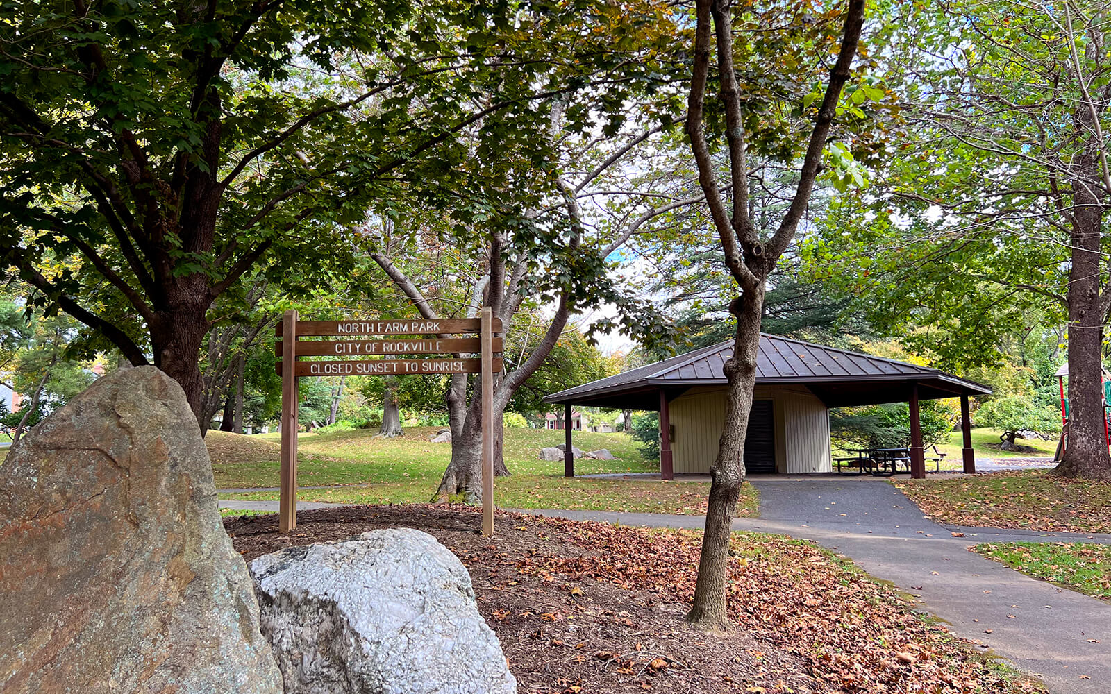 A wooden park sign and large rocks stand near a paved path leading to a covered pavilion surrounded by trees in North Farm Park.