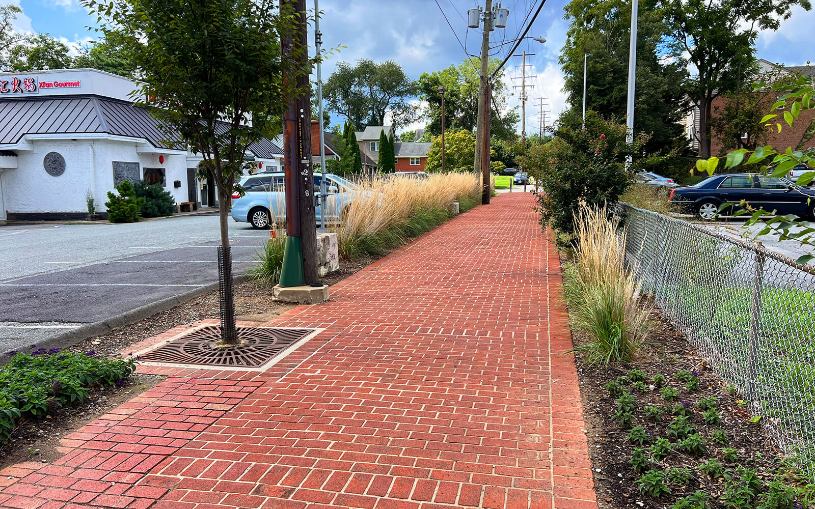 A red brick sidewalk runs alongside a chain-link fence and landscaped plants, with a restaurant and utility poles visible in the background.