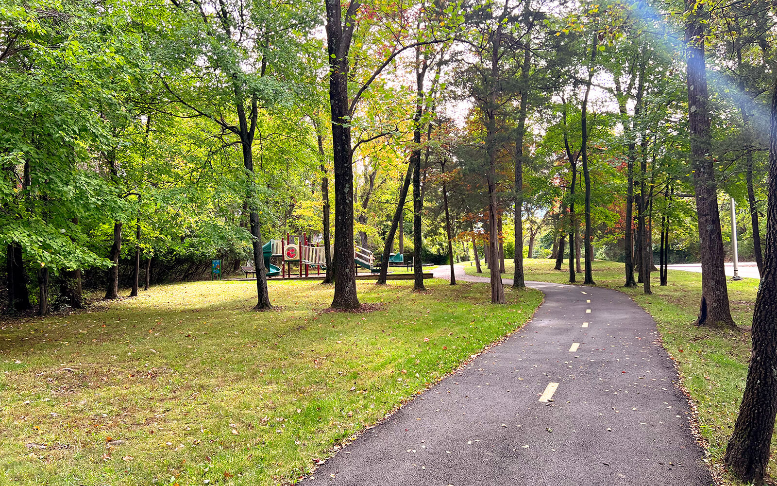 A paved walking path curves through a park with green grass, trees, and a playground visible in the background.