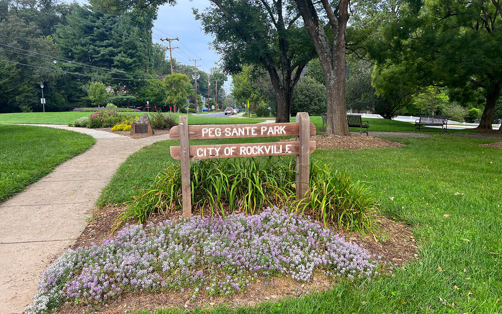 A wooden sign reading “PEG SANTE PARK CITY OF ROCKVILLE” stands among flowers and grass, with a sidewalk and trees in the background.