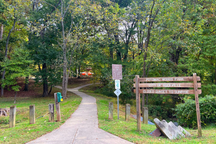A paved path leads into Potomac Woods Park, marked by a wooden sign and surrounded by trees. Signs indicate park hours and rules.