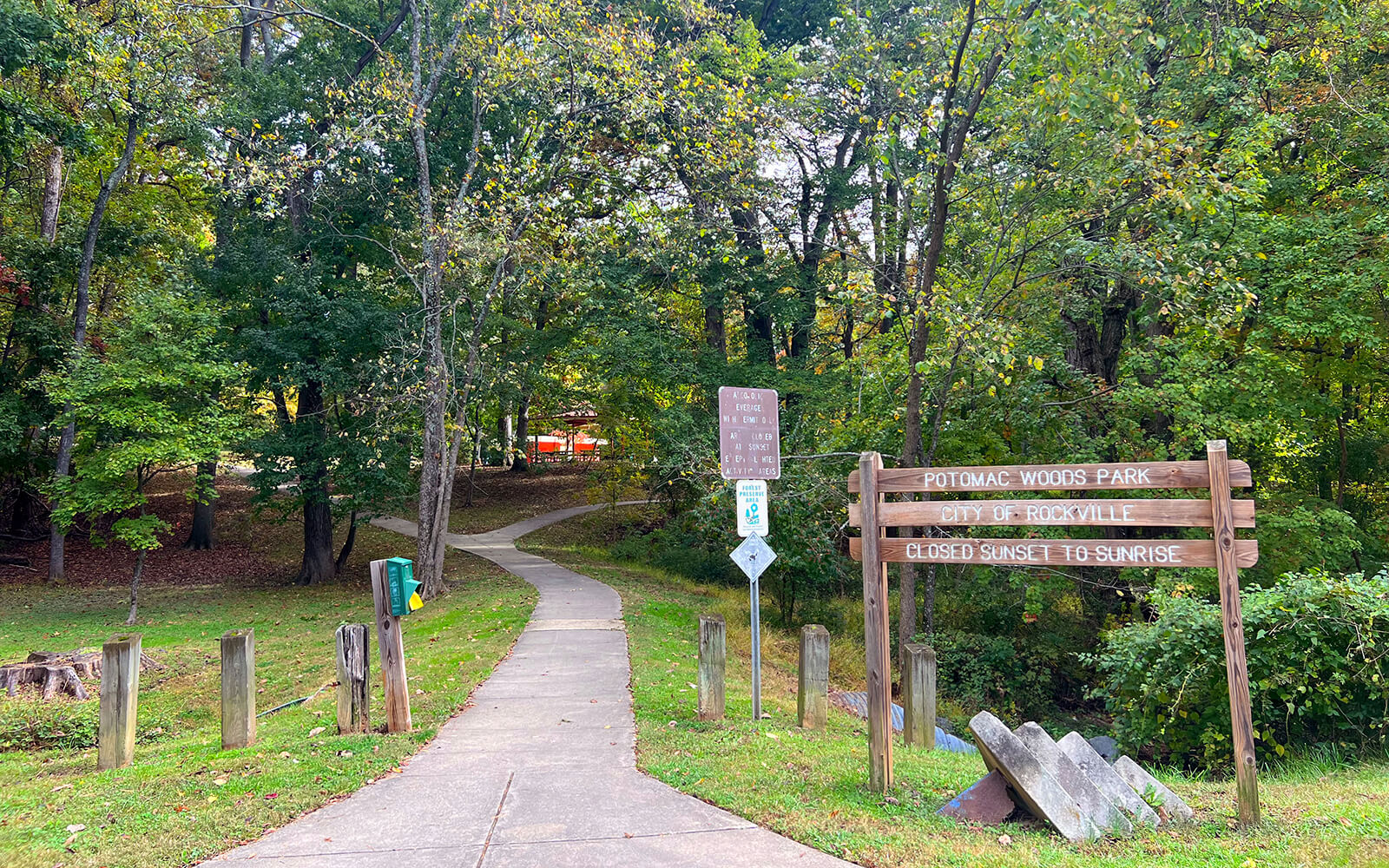 A paved path leads into Potomac Woods Park, marked by a wooden sign and surrounded by trees. Signs indicate park hours and rules.
