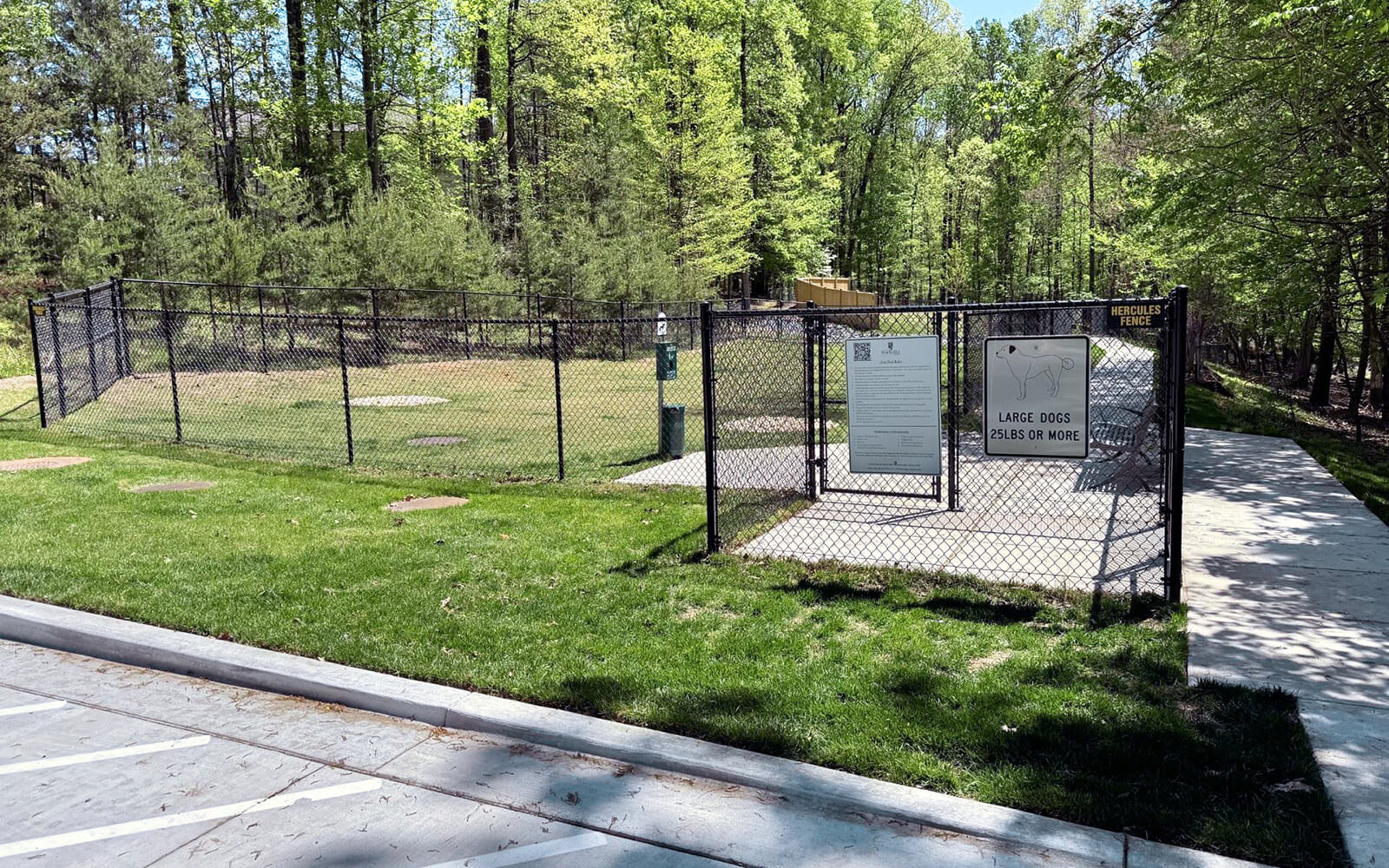 A fenced dog park area with a sign for large dogs, surrounded by grass and trees, with a sidewalk leading to the entrance.