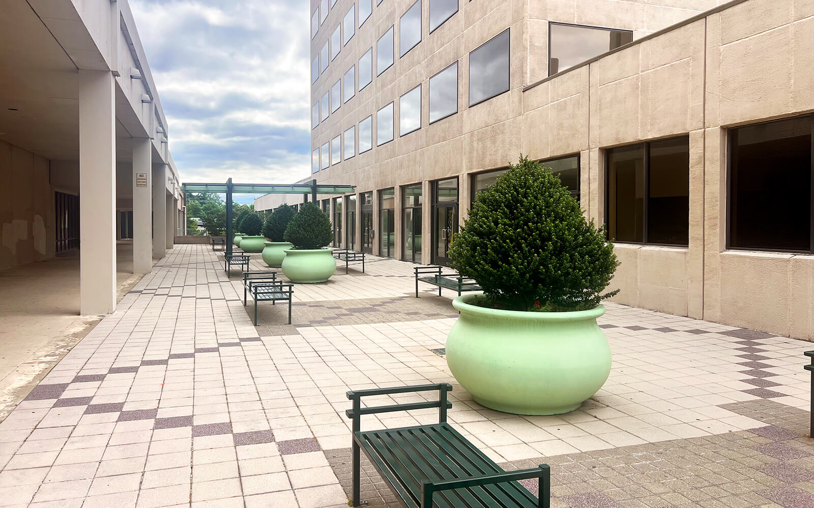 Outdoor courtyard with large green planters, small trees, metal benches, and a building with many windows under a cloudy sky.
