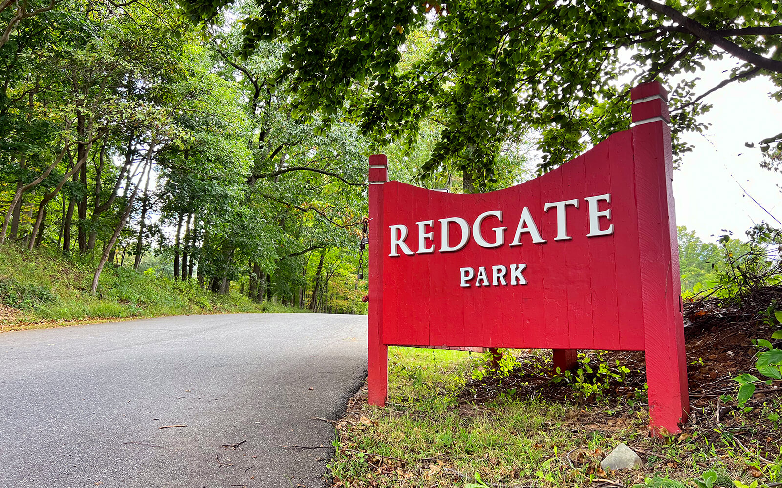 A red sign reading "Redgate Park" stands beside a paved path surrounded by green trees and grass.