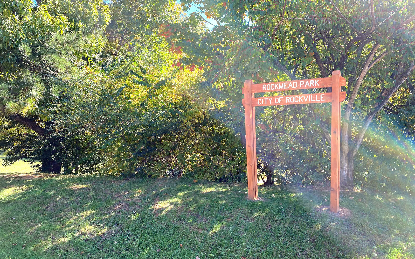 A wooden sign reading "Rockmead Park, City of Rockville" stands on a grassy area in front of trees under bright sunlight.