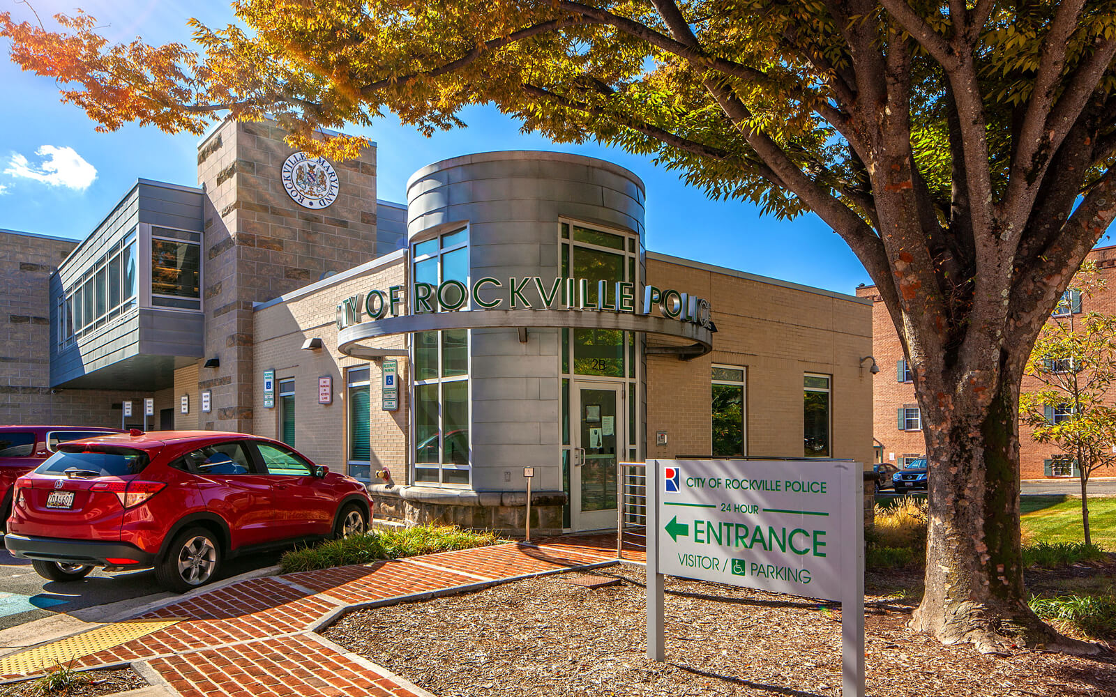 Modern building with large windows and "Rockville Police" sign; a red car is parked outside, and a sign directs visitors to the entrance and parking area.