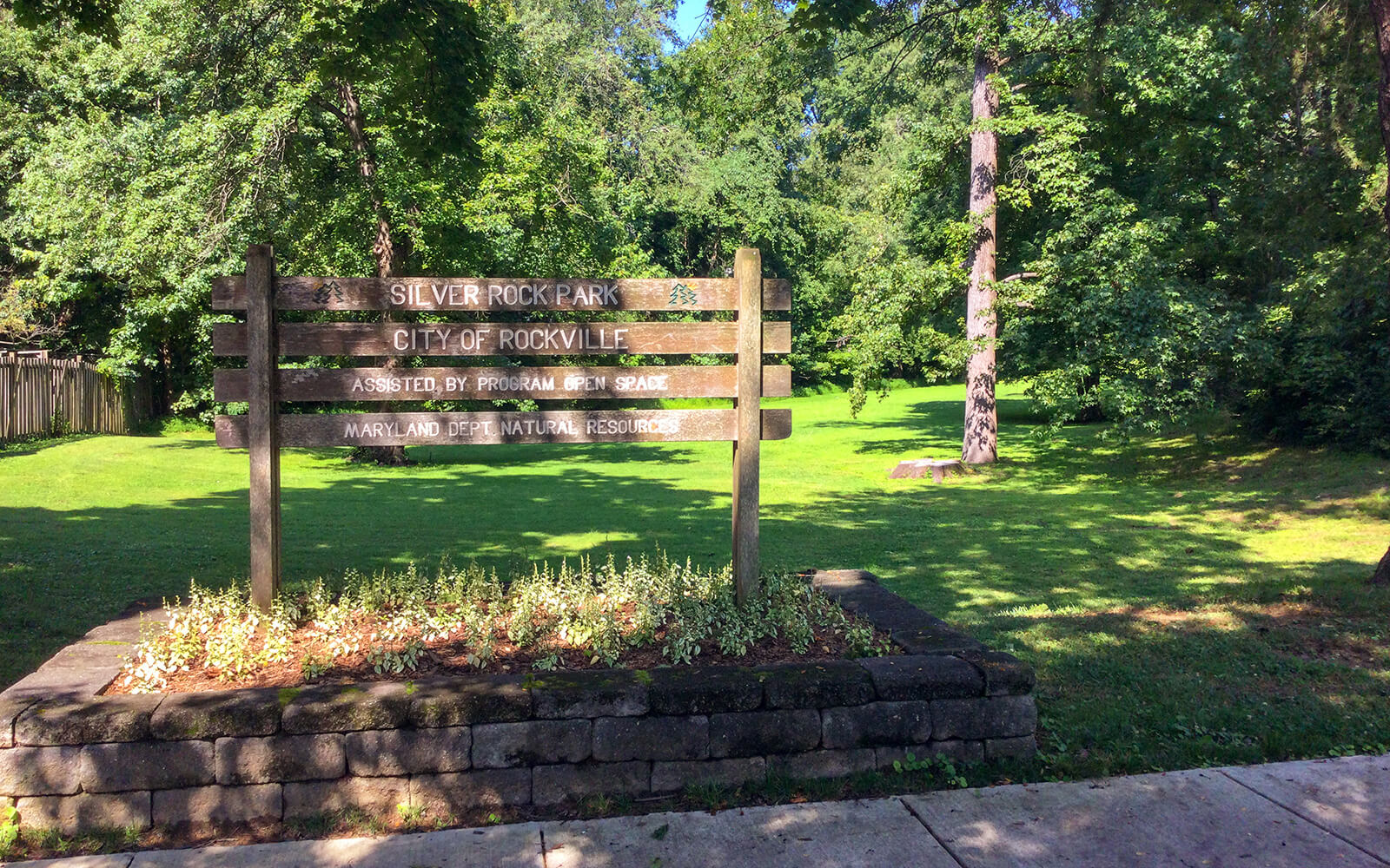 A wooden sign for Silver Rock Park, City of Rockville, stands on a stone base in a grassy area surrounded by trees.