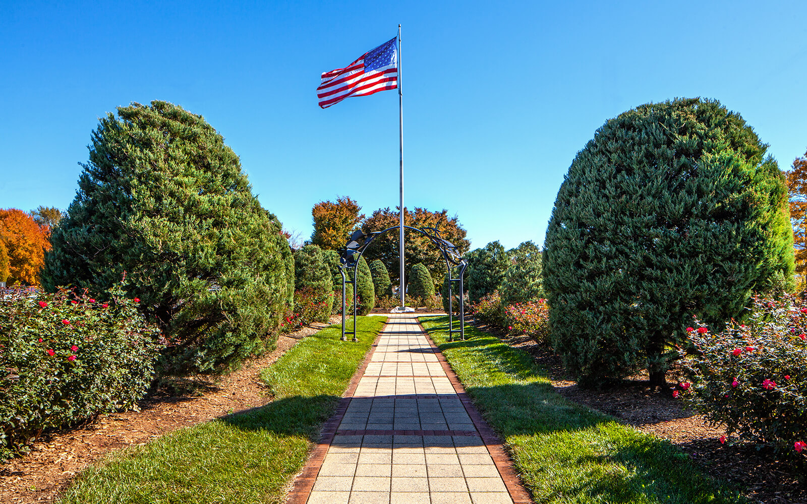 A paved walkway lined with trimmed bushes leads to an American flag on a flagpole under a clear blue sky.