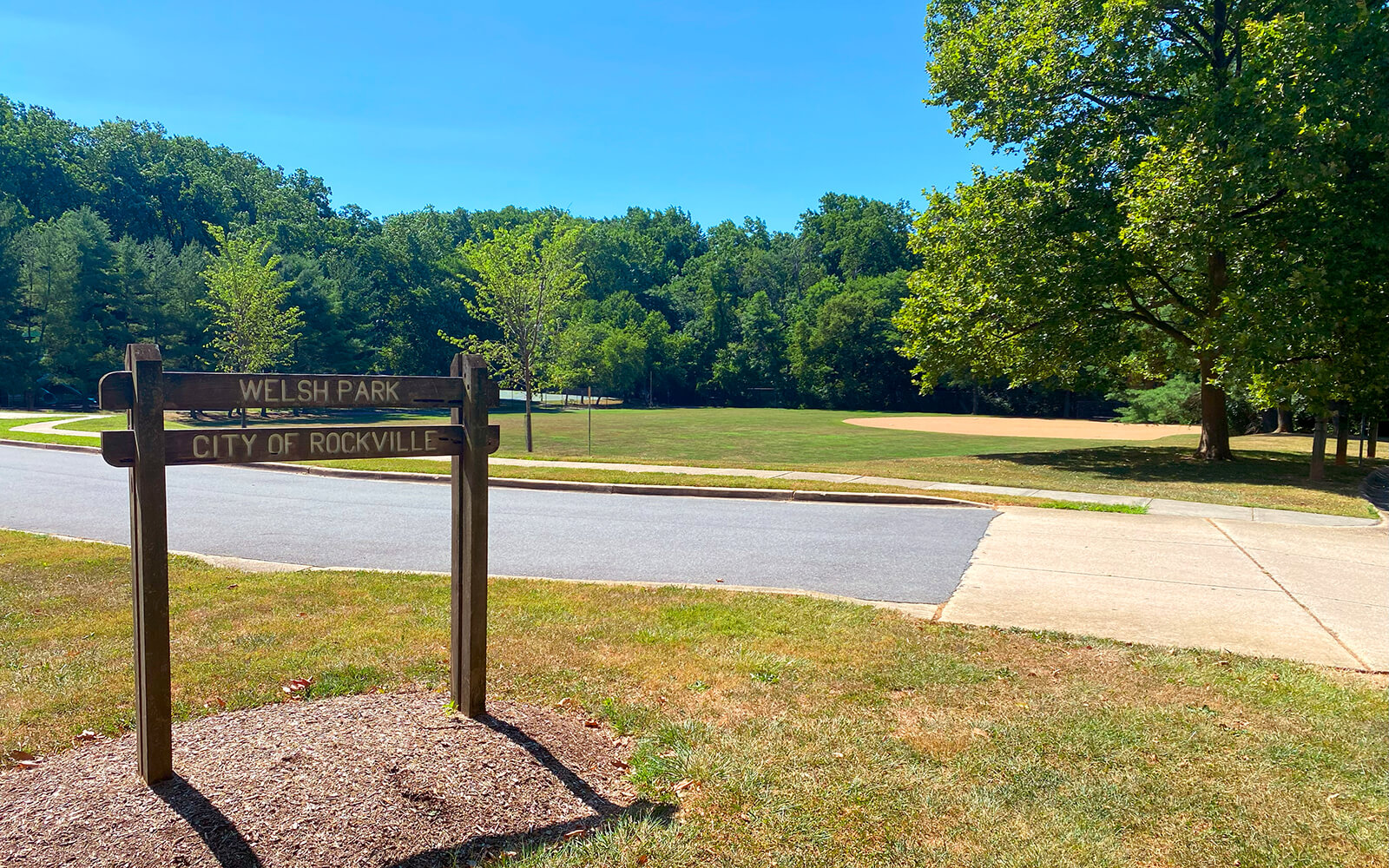 A wooden sign reads "Welsh Park City of Rockville" in front of a grassy park area with trees and a clear blue sky.