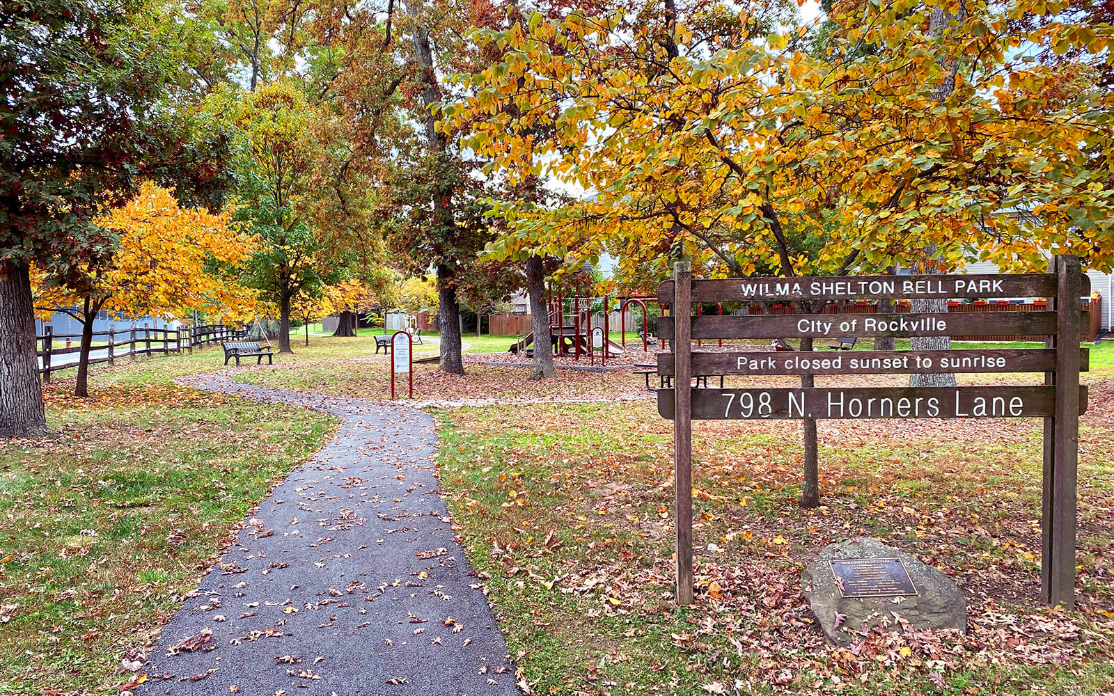 Paved path through Wilma Shelton Bell Park with autumn foliage, benches, and a wooden sign displaying park information and address.