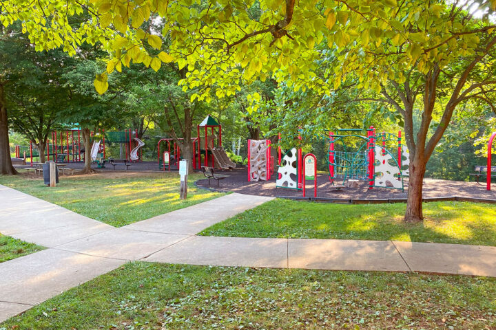 Playground with slides and climbing equipment surrounded by trees and grass, bordered by concrete walkways on a sunny day.