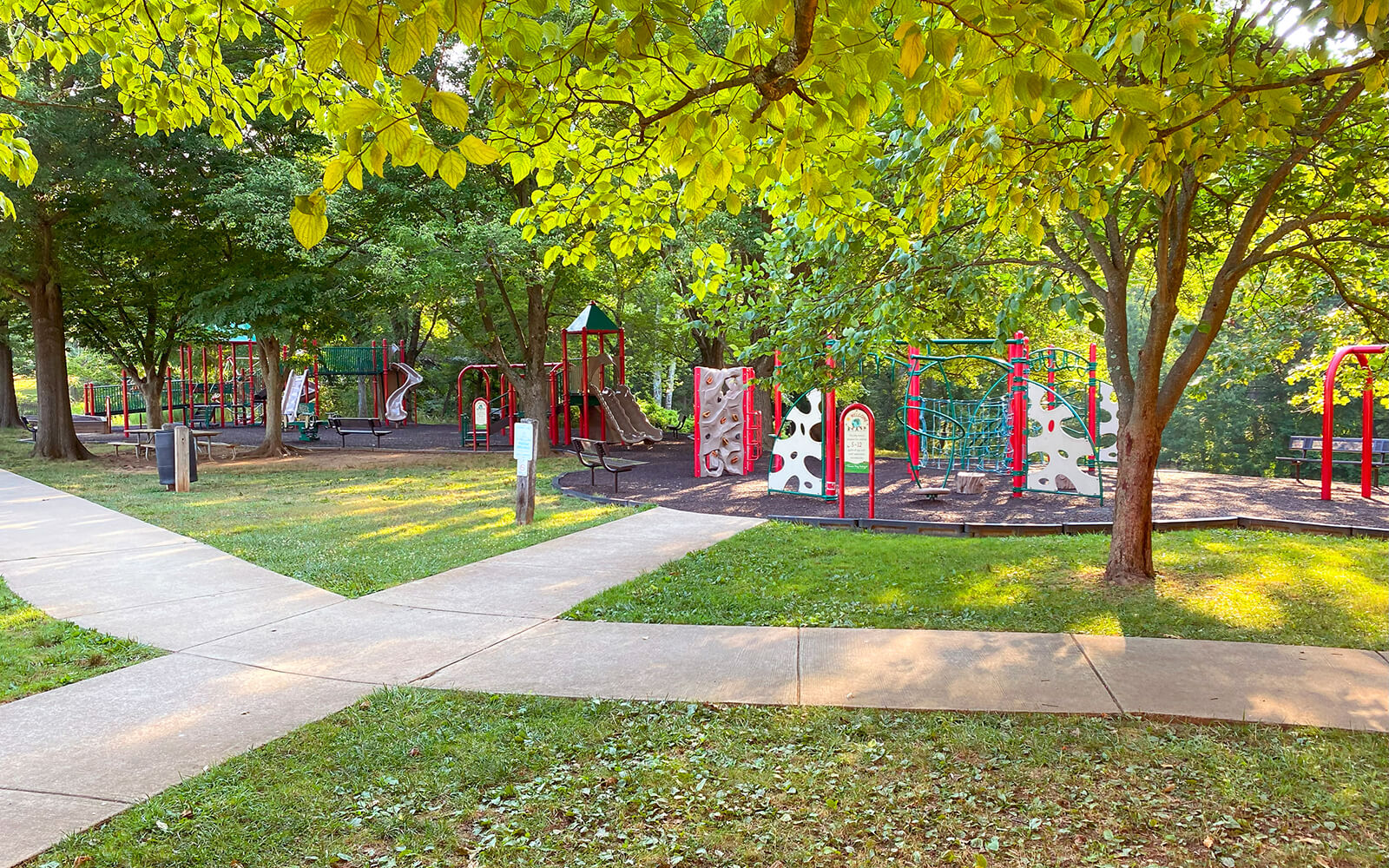 Playground with slides and climbing equipment surrounded by trees and grass, bordered by concrete walkways on a sunny day.