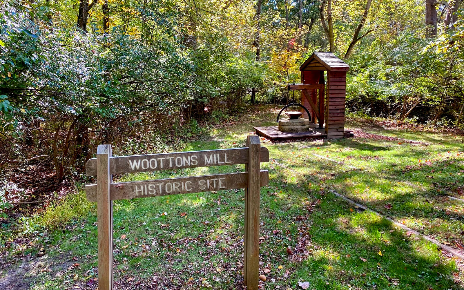 Wooden sign reading "Woottons Mill Historic Site" in front of an old mill structure surrounded by trees and grass in a wooded area.