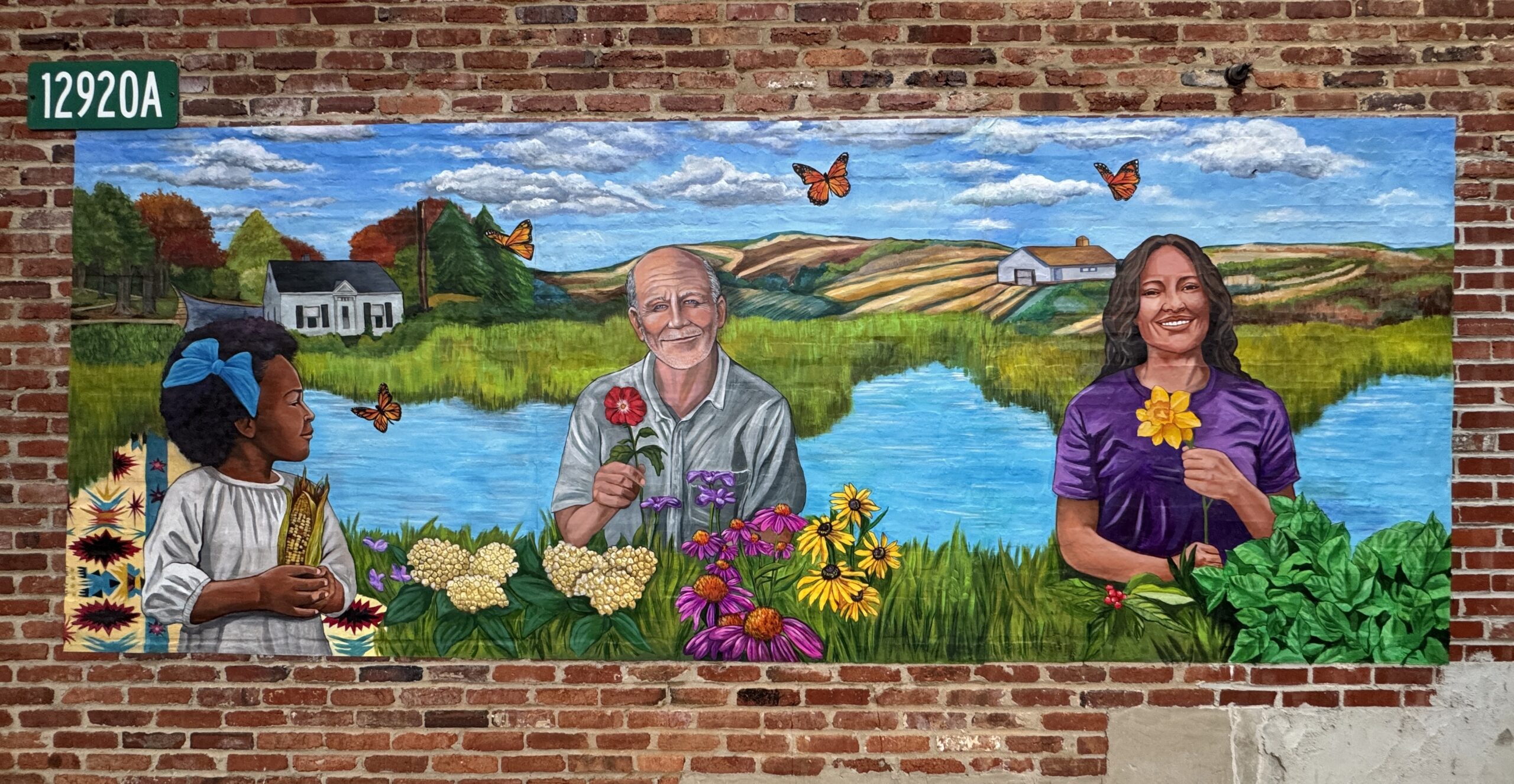A mural on a brick wall shows a young girl, an older man, and a woman holding flowers in front of a lake and hills, with butterflies flying in the sky.