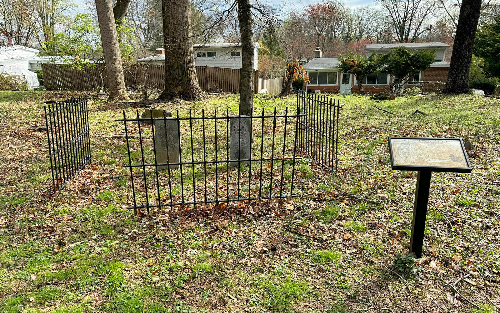 A small fenced grave site with two headstones stands in a grassy yard near trees and residential houses; an informational plaque is nearby.