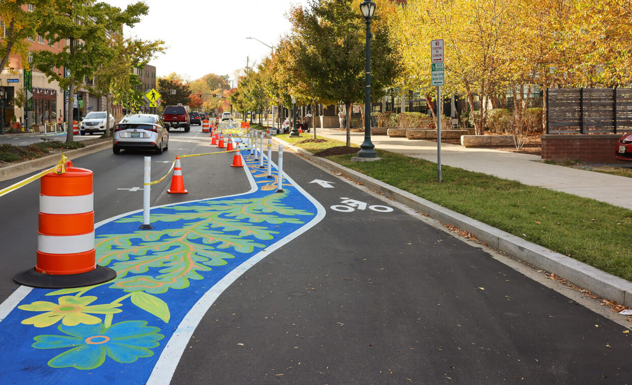 A street with a bike lane marked by poles, orange traffic cones, and decorative blue and green leaf patterns; cars and trees line the road.