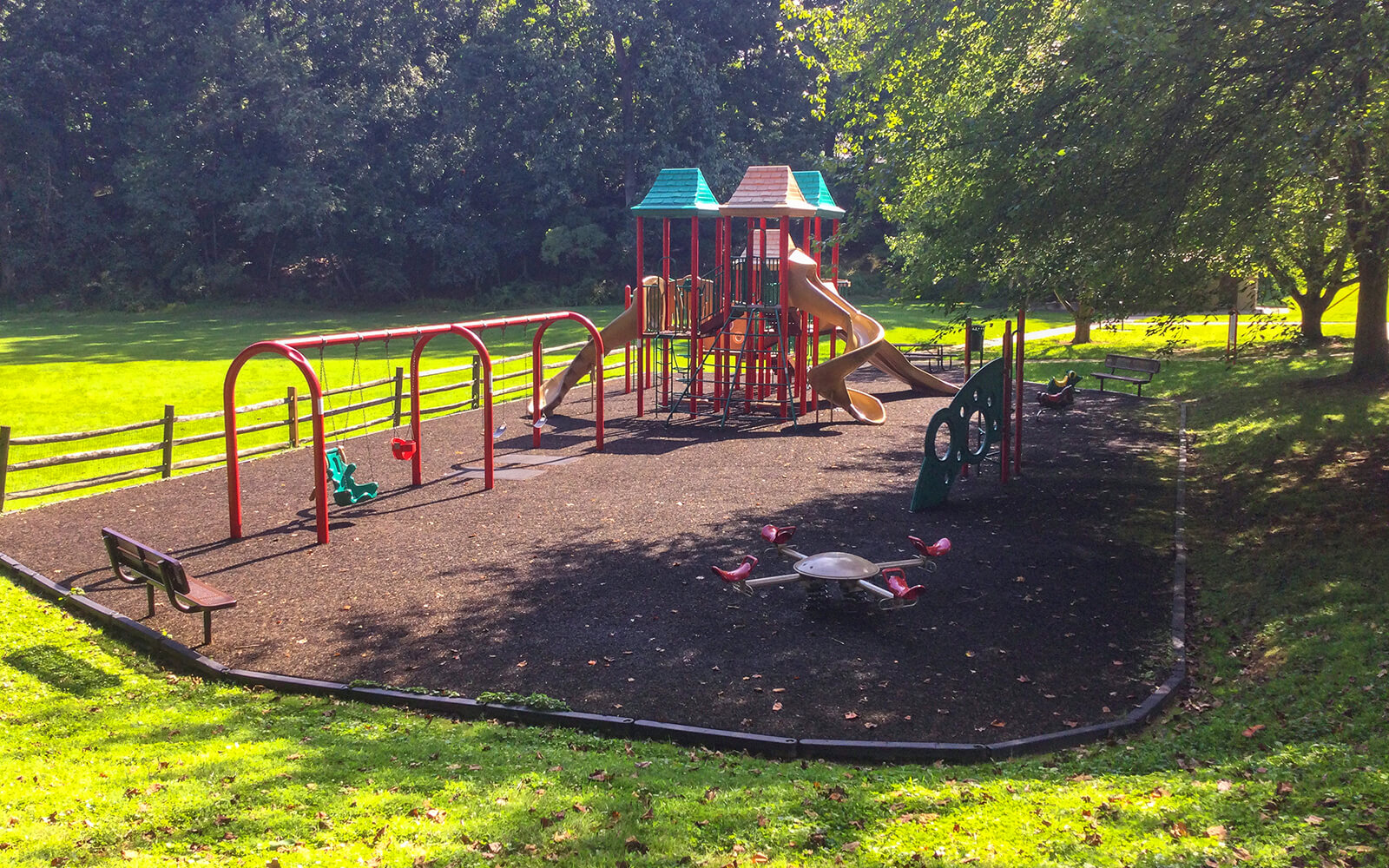 A playground with swings, slides, climbing structures, and a roundabout, surrounded by grass and trees on a sunny day.