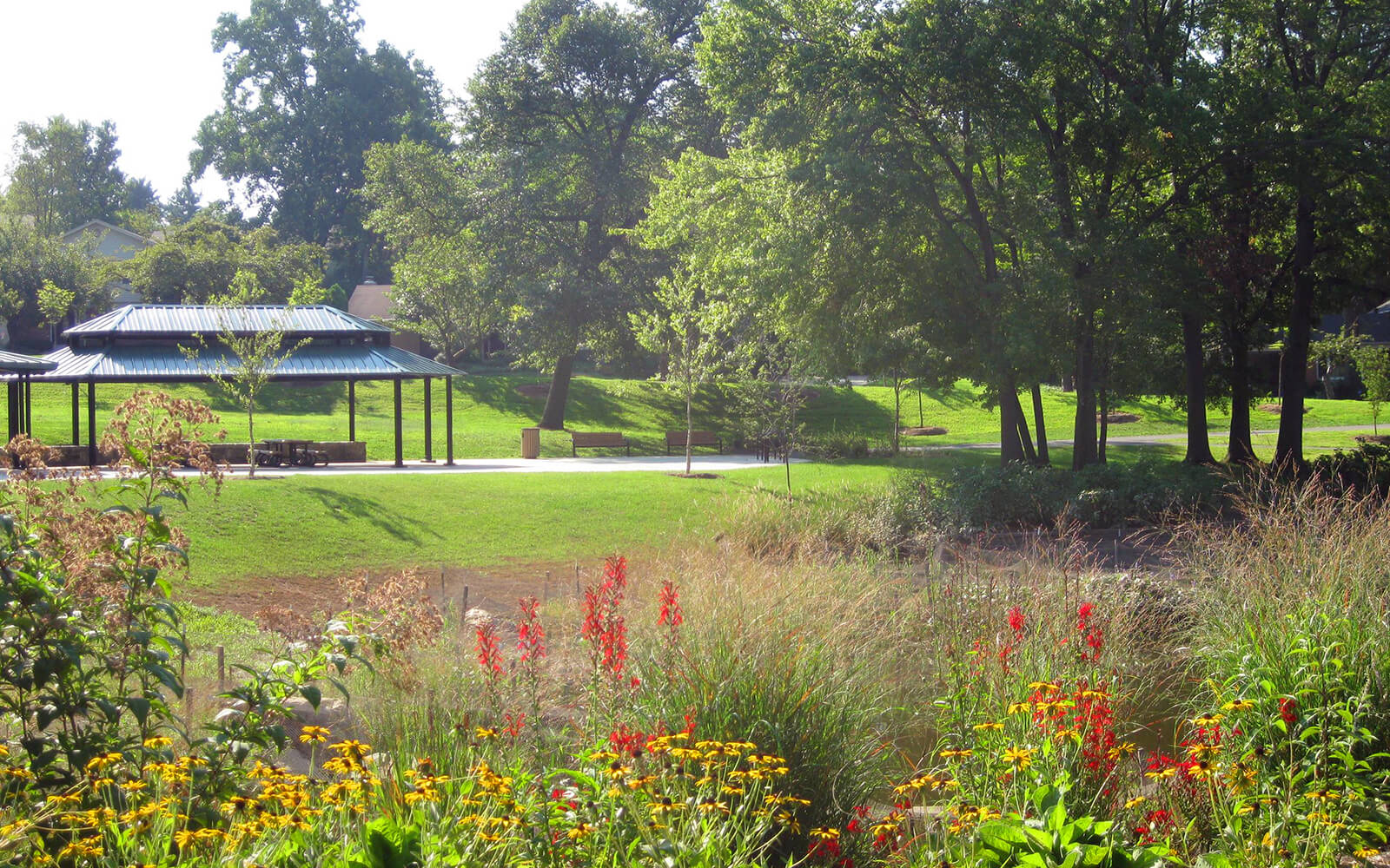 A park scene with a pavilion, picnic tables, grassy lawns, trees, and colorful flowers in the foreground on a sunny day.